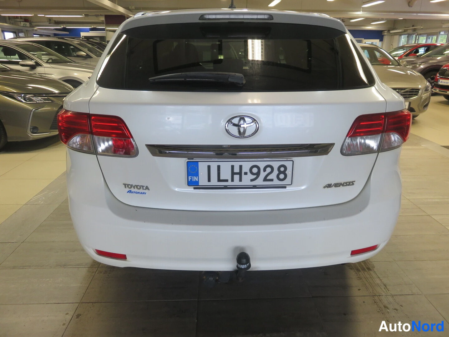 Takaa: Valkoisen farmariauton takaosa. Perällä punaiset takavalot, Toyota-merkki, Avensis-teksti ja nokkakoukku. Rekisteritunnus.
