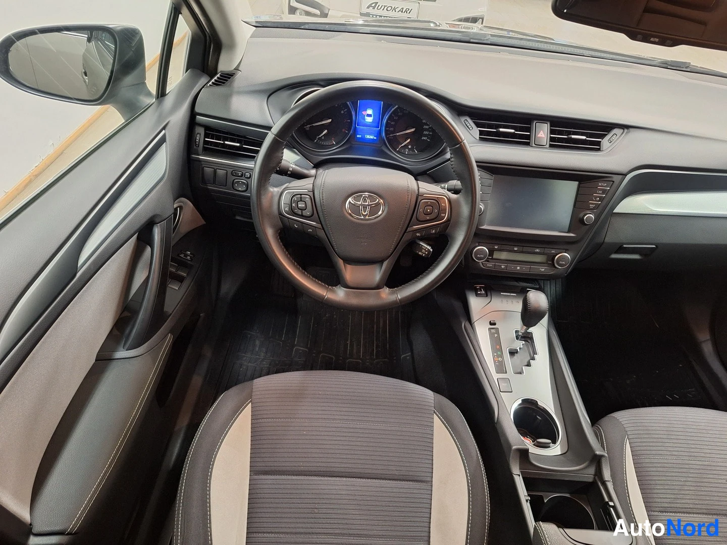 Edestä: Kuljettajan puoleinen sisätilanäkymä: Toyota-ohjauspyörä, mittaristo, automaattivaihteen valitsin ja infotainment-näyttö.