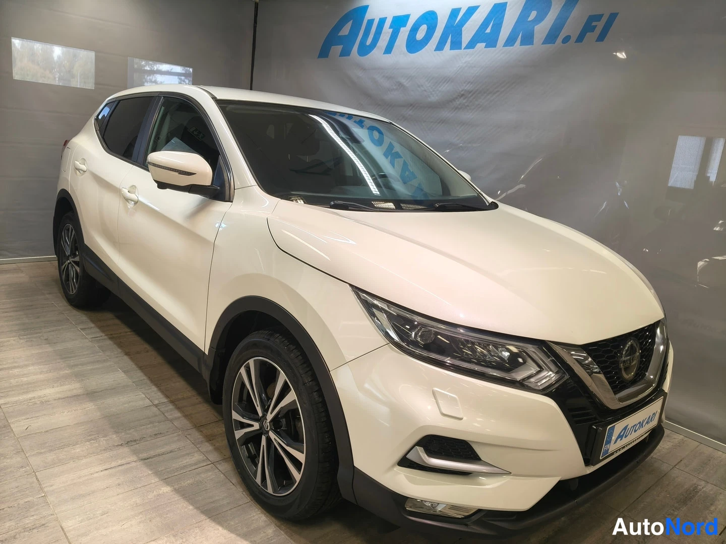 Etuviisto, nokka oikealle: Nissan Qashqai valkoinen kulmikkaasti kuvattu etuosasta oikealta showroomissa, LED-ajovalot ja monihaarapeili pyörät näkyvät.