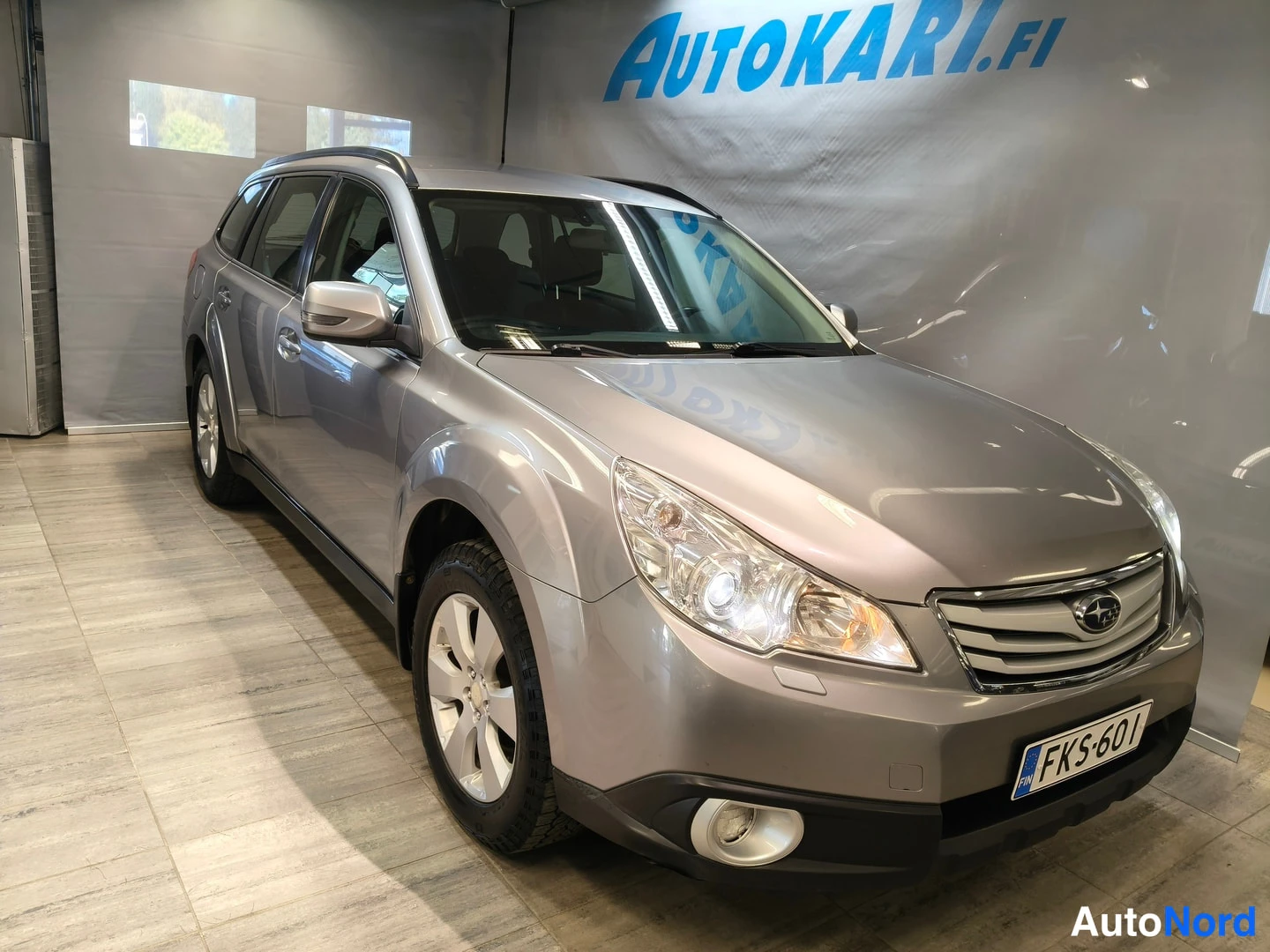 Etuviisto, nokka oikealle: Harmaa Subaru Outback -farmariAutoKari.f-taulun edessä myyntihallissa, etunäkymä vasemmasta kolmasosakulmasta, rekisteritunnus.