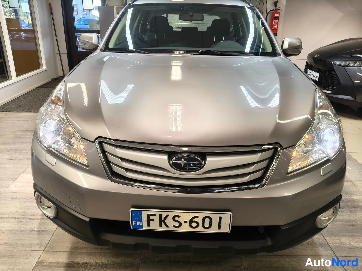 Edestä: Harmaa Subaru Outback suoraan edestä kuvattuna, moottoritilan konepelti ja Subaru-merkki näkyvät, rekisteritunnus.