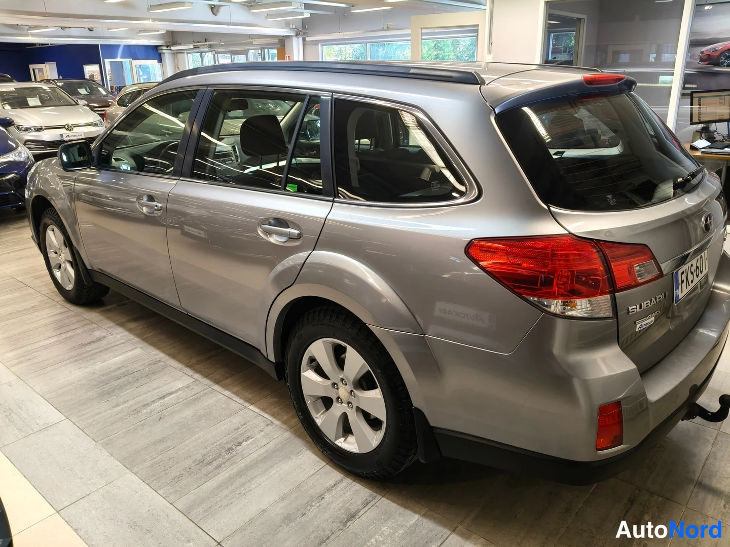 Takaviisto: Harmaa Subaru Outback -farmari takavasemmalta kuvattuna autosalissa, takavalot ja avattu takaluukku näkyvissä, rekisteritunnus.