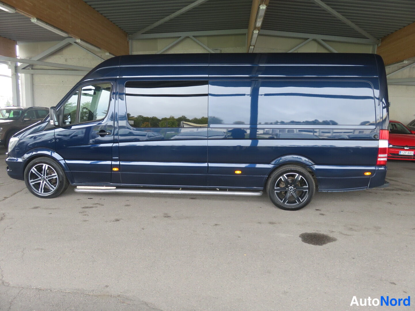 Sivulta: Pakkasen sininen pitkä Mercedes-Benz Sprinter -pakettiauto sivusuunnassa autotallin sisällä, isoissa vanteissa ja sivulaatikoilla.