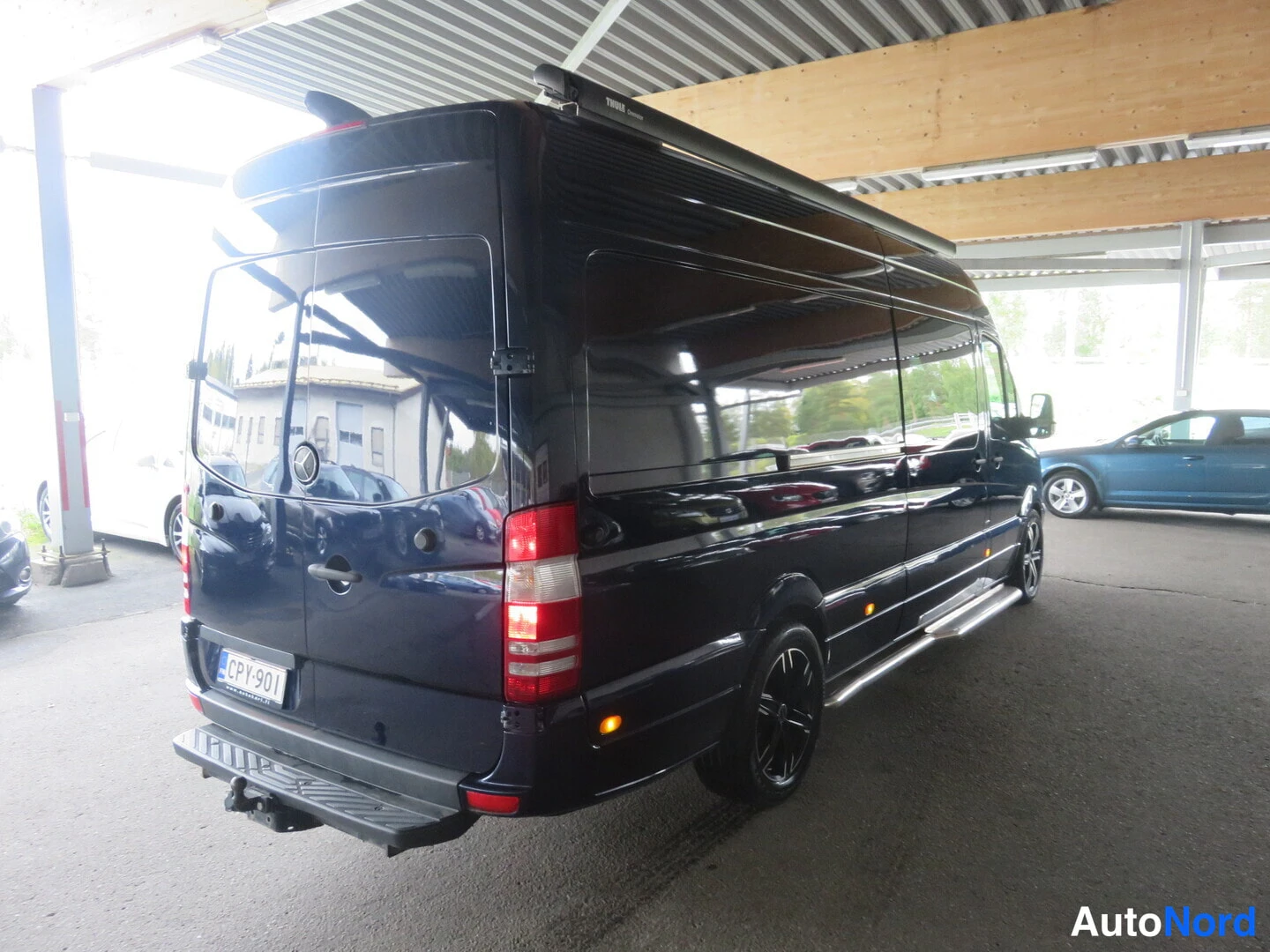 Takaviisto: Tummansininen Mercedes-Benz -pakettiauto takaviistosta oikealta; rekisteri, mustat vanteet, kromilistoja ja takaossaan korokkeella.