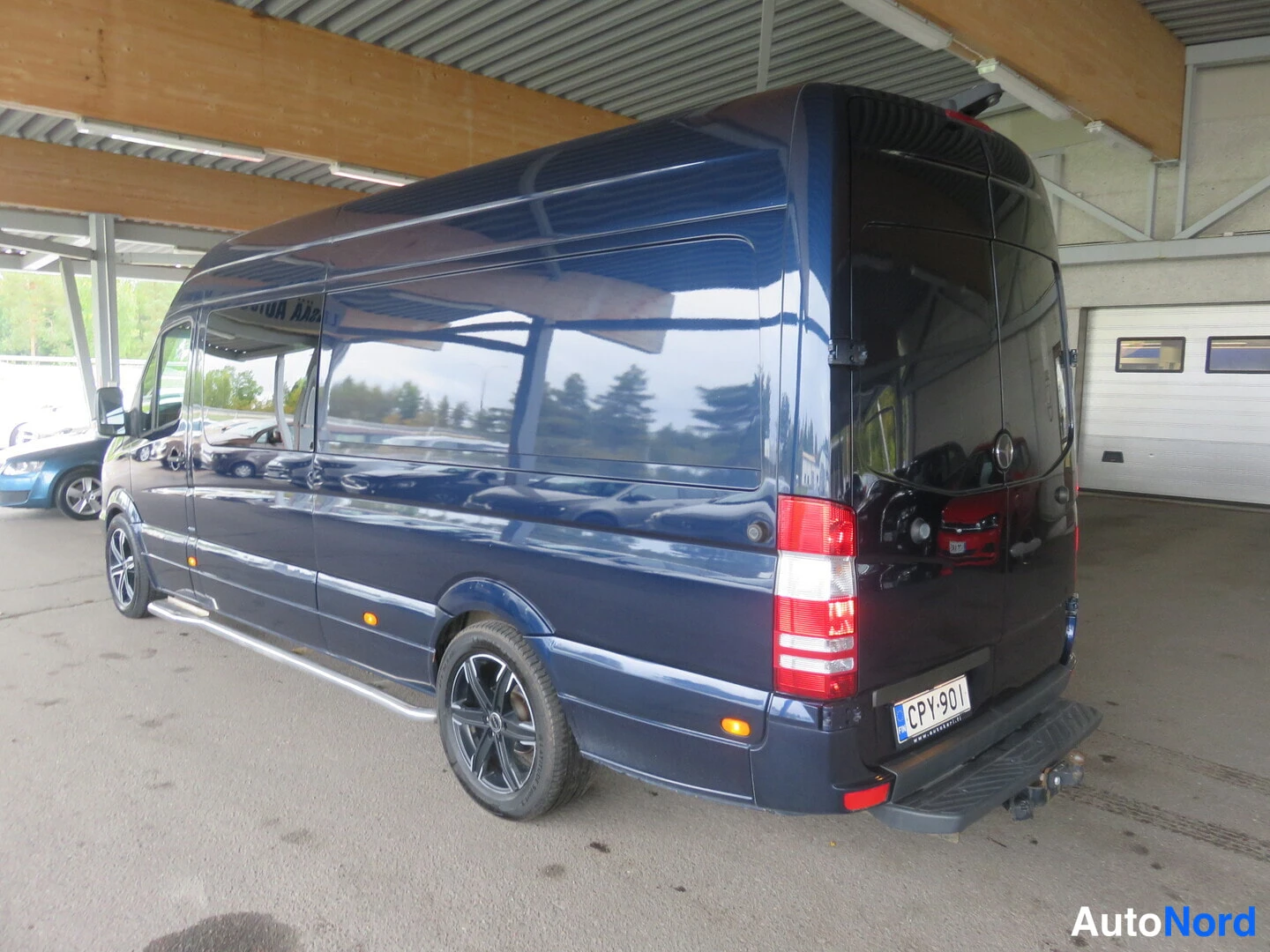 Takaviisto: Tummansininen Mercedes-Benz -pakettiauto takaviistosta vasemmalta; rekisteri, mustat vanteet, kromilista, taakse kytketty vetokoukku.