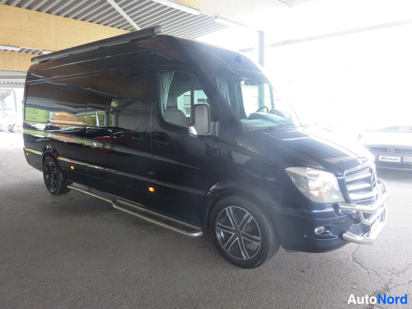 Etuviisto, nokka oikealle: Tummansininen Mercedes-Benz Sprinter -pakettiauto etuviistöstä autokatoksessa, kylki ja konepelti oikealla, auton kyljessä ikkunat.