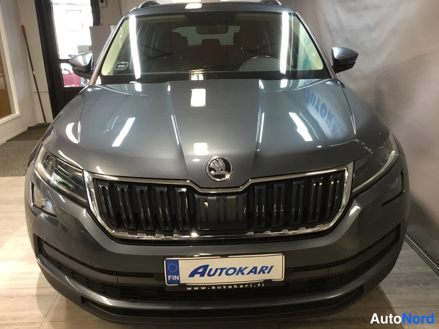 Edestä: Harmaanvärinen Škoda Kodiaq etunäkymästä autonäyttelytilassa, etusäleikkö ja ajovalot esillä.