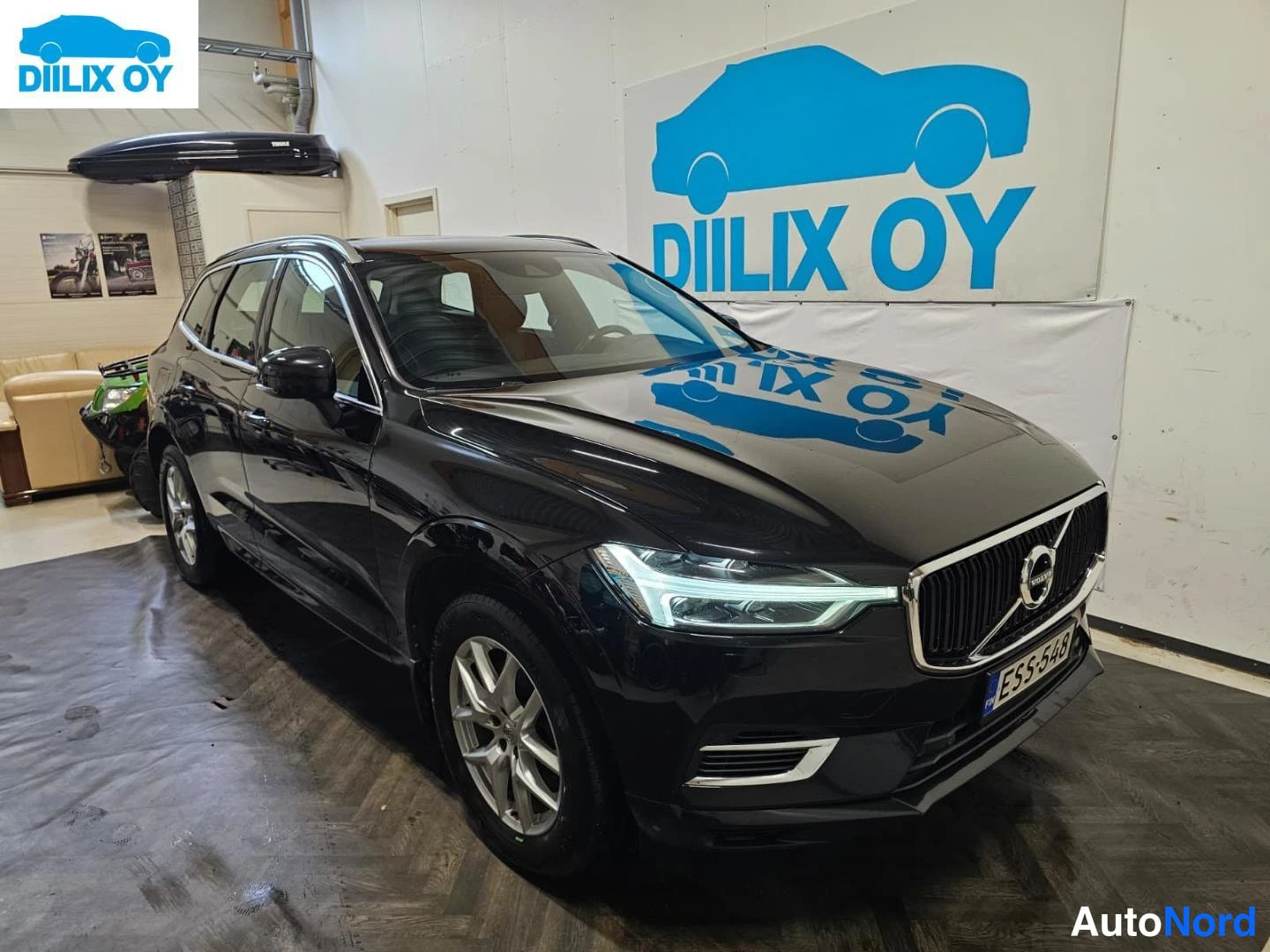 Etuviisto, nokka oikealle: Musta Volvo XC60 -farmarikori etukulmasta sisätiloissa, rekisterikilpi.