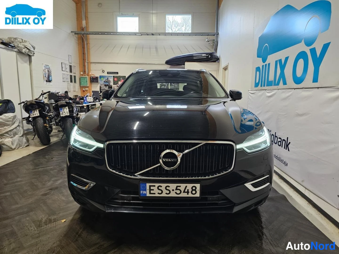 Edestä: Musta henkilöauto suoraan edestäpäin, etusäleikkö ja ajovalot näkyvissä, rekisteritunnus.