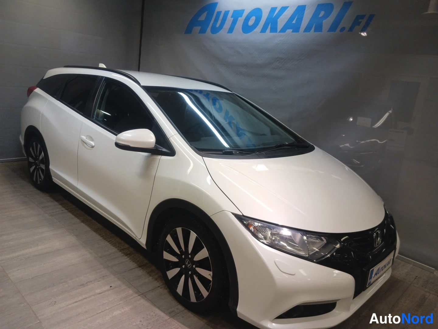 Etuviisto, nokka oikealle: Valkoinen Honda Civic Tourer -farmari näkyvissä etukulmasta myymälän seinan edessä.