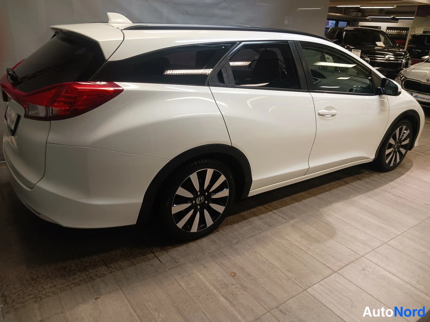 Takaviisto: Valkoinen Honda Civic Tourer -farmarivauto sivulta takaa kuvattuna autoshallissa, mustat pölykkeet ja kevytmetallivanteet näkyvissä.