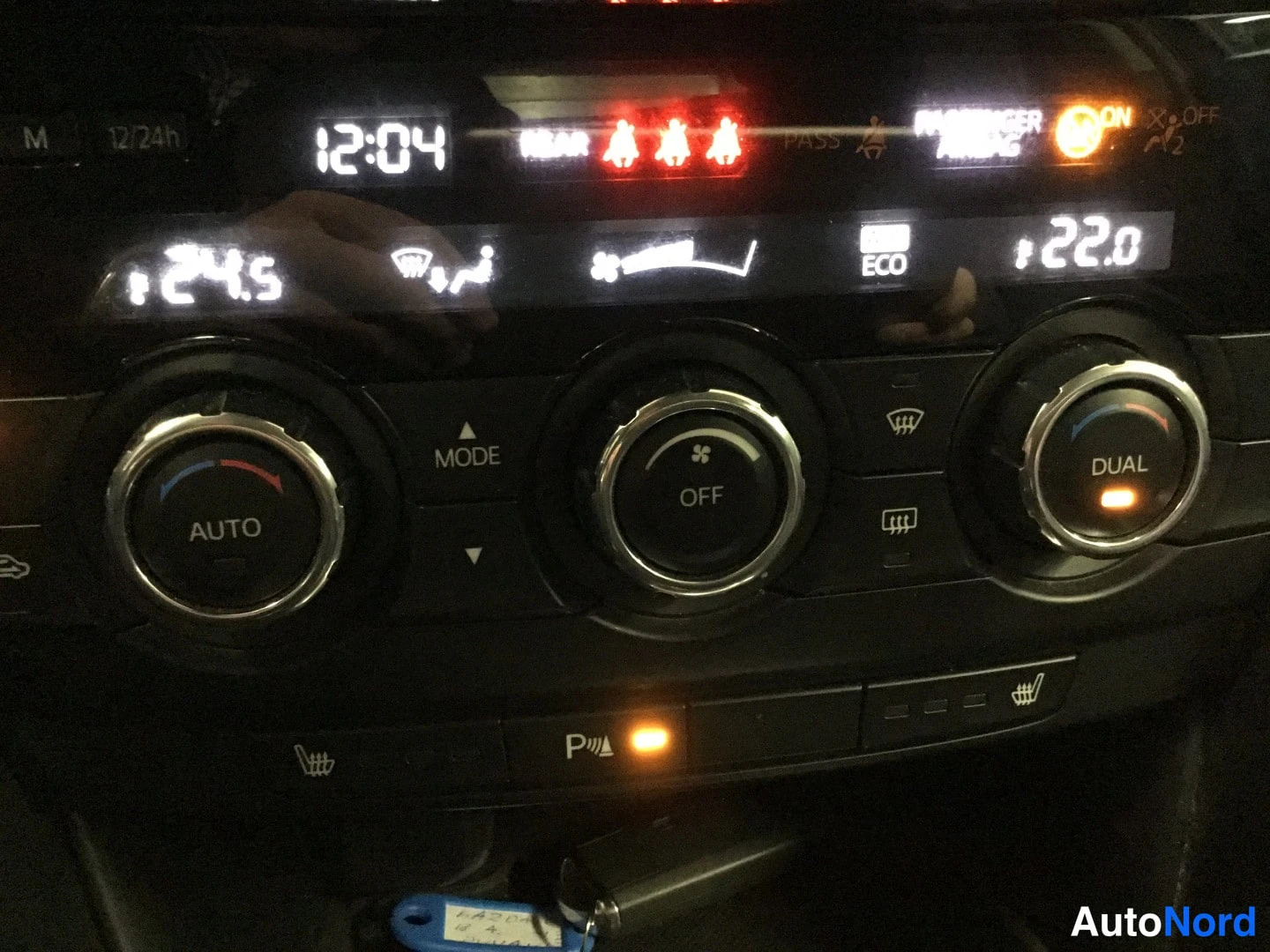 Muu: Auton ilmastointipaneeli, näytössä ajat 12:04 ja 24,5 / 22,0, nappuloilla AUTO ja DUAL, alaohjaus lamppu merkinnällä.