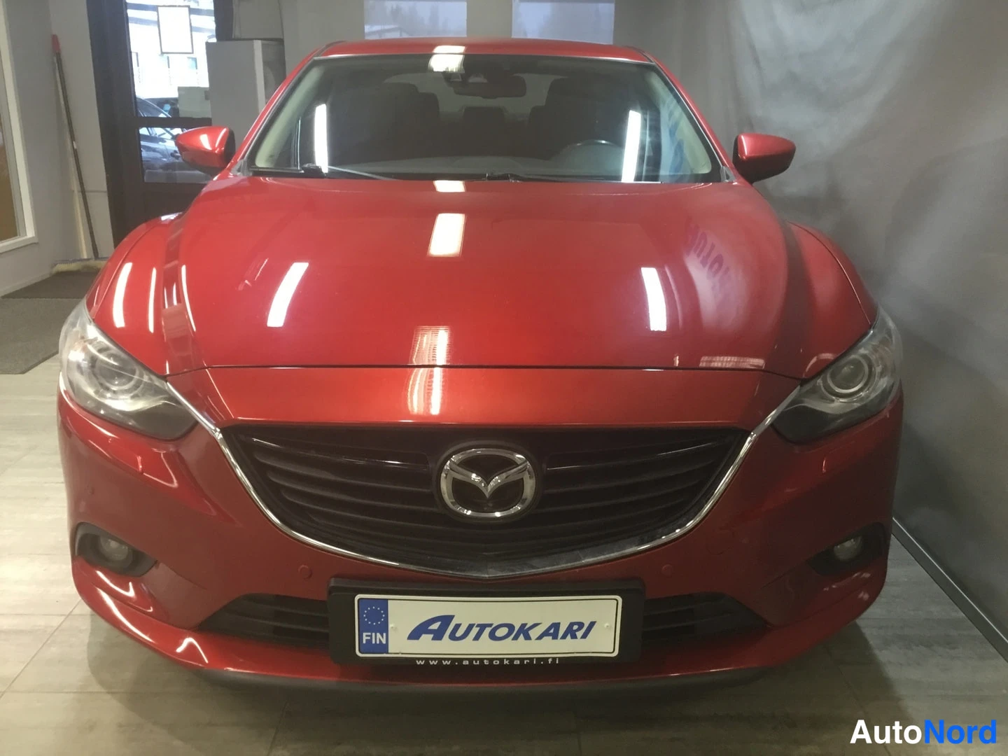 Edestä: Punaisen Mazda 6:n etupää suoraan edestä kuvattuna nähtävissä Mazda-merkki, etugrilli, ajovalot ja Autokari-kilpi.