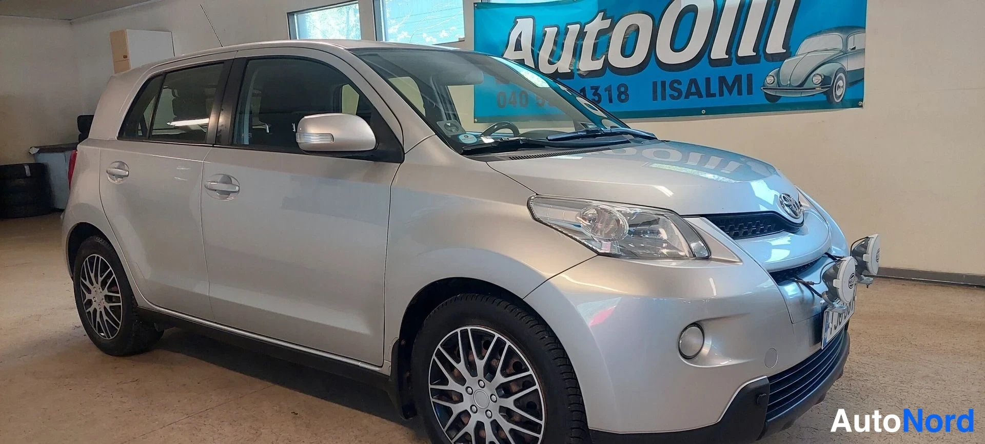 Etuviisto, nokka oikealle: Hopeanharmaa Toyota Urban Cruiser etukulmasta oikealta kuvattuna hallissa; keulan grilli, T-merkki ja etupuskurin lisävalot ovat selvästi näkyvissä.