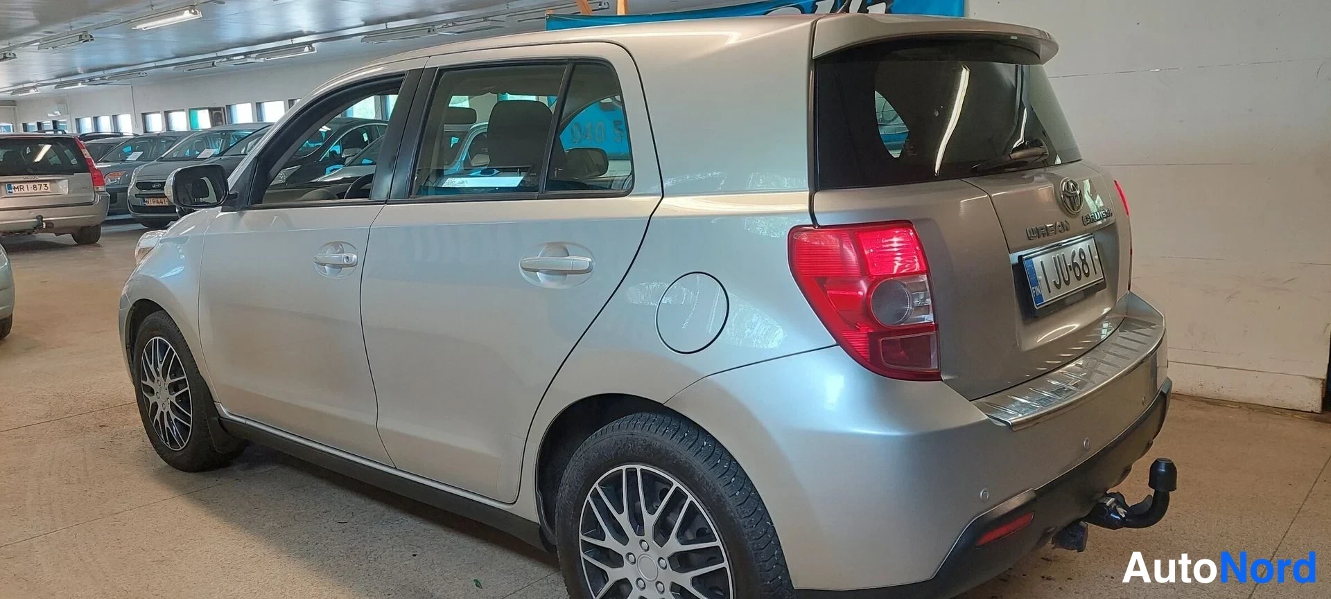 Takaviisto: Hopeanharmaa Toyota Urban Cruiser takakulmasta vasemmalta kuvattuna autovahviihallissa; takavalo, rekisterikilpi J UJ-681 ja takavarrassa oleva vetokoukku ovat