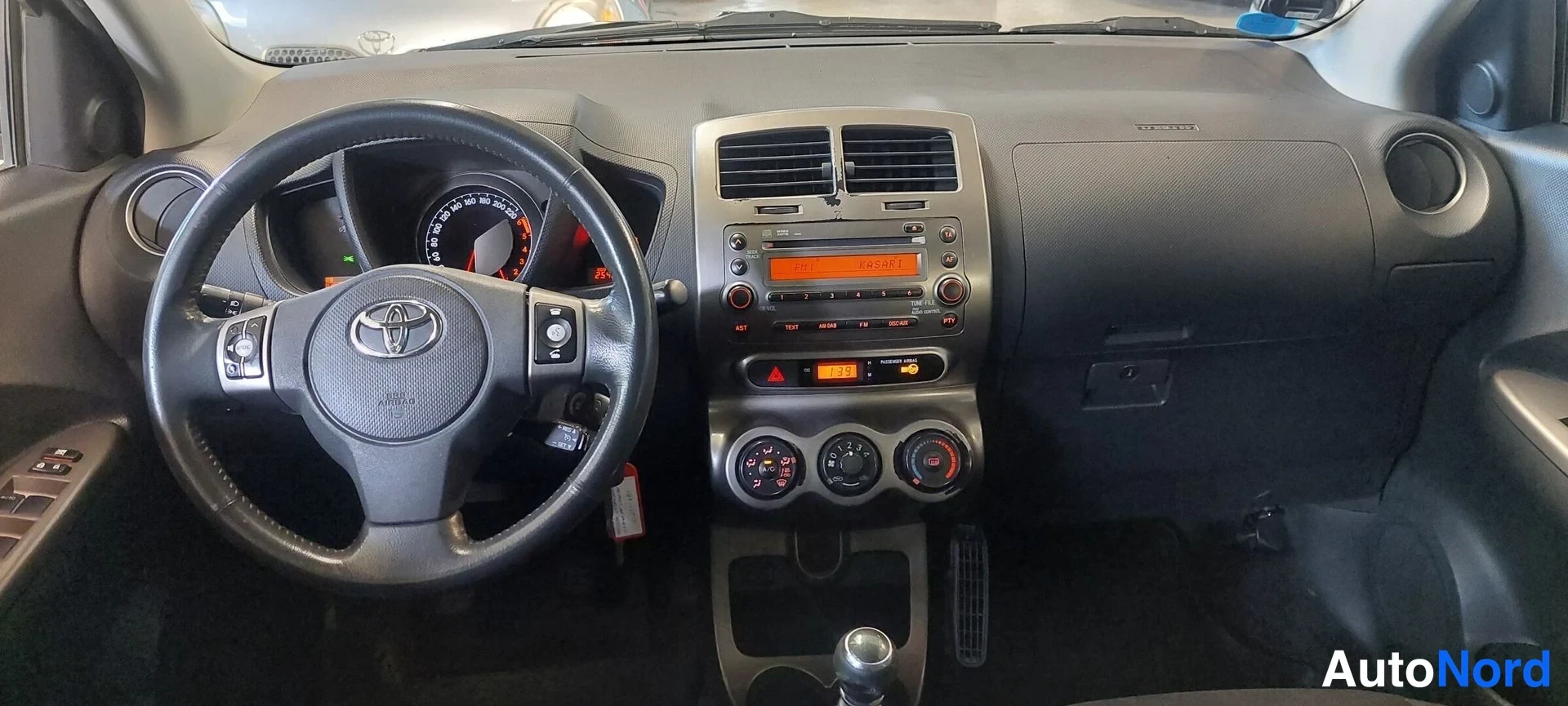 Muu: Ohjaamo edestä: Toyota-ohjauspyörä, kojelauta, radiopaneeli, manuaalivaihteen keila ja polkimet näkyvät.