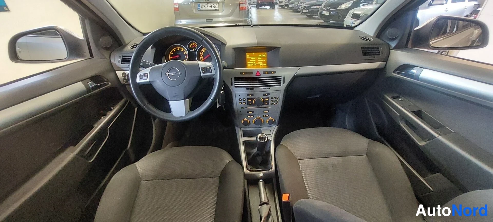 Muu: Auton etuistuimilta kuvattu ohjaamo: Opel-ohjauspyörä, mittaristo, keskikonsoli, manuaalivaihdevipu ja tummat kangasistuimet.