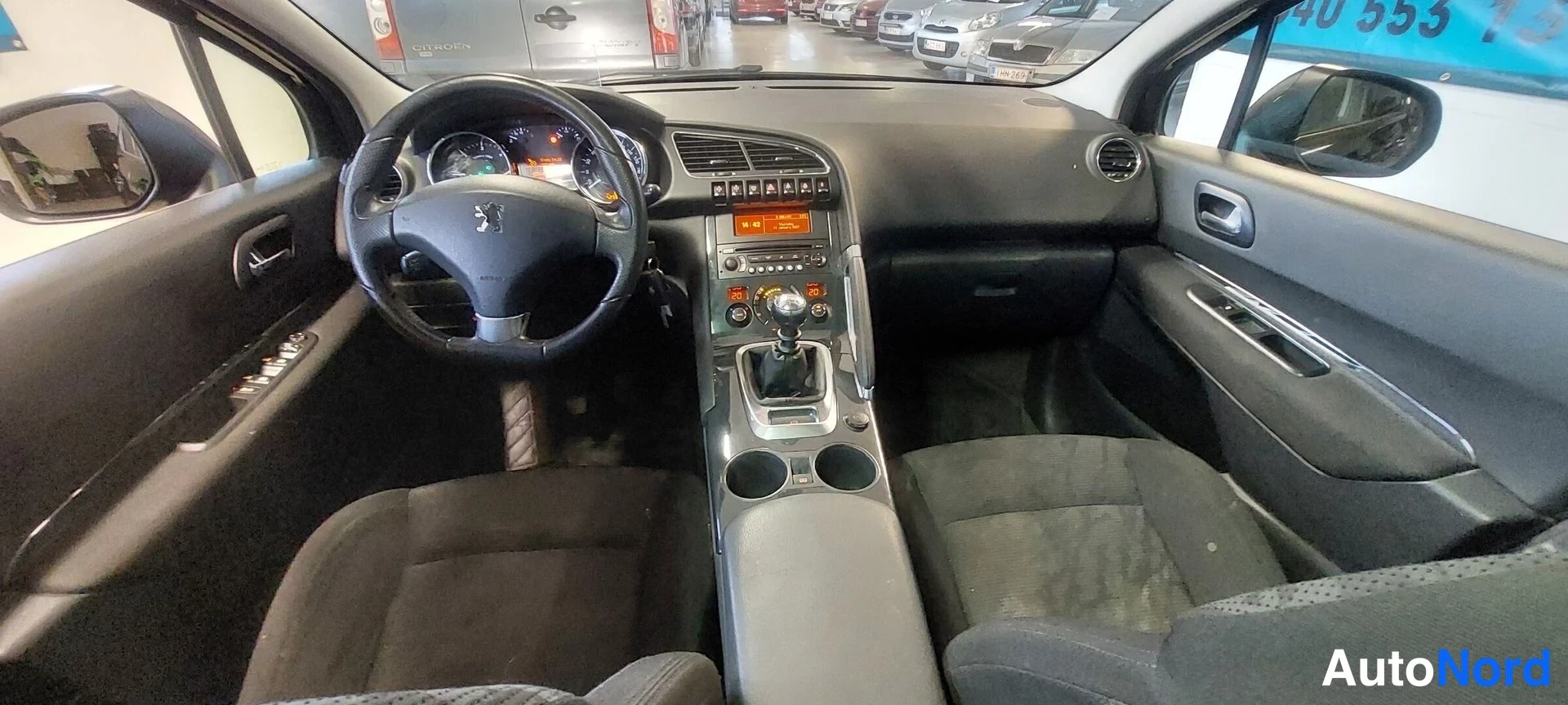 Muu: Auton sisätila näkyy takaistuimelta: tumma kojelauta, ohjauspyörä Peugeot-embleemillä, manuaalivaihteenvaihtaja ja kaksi etuistuinta kangasverhoilulla.