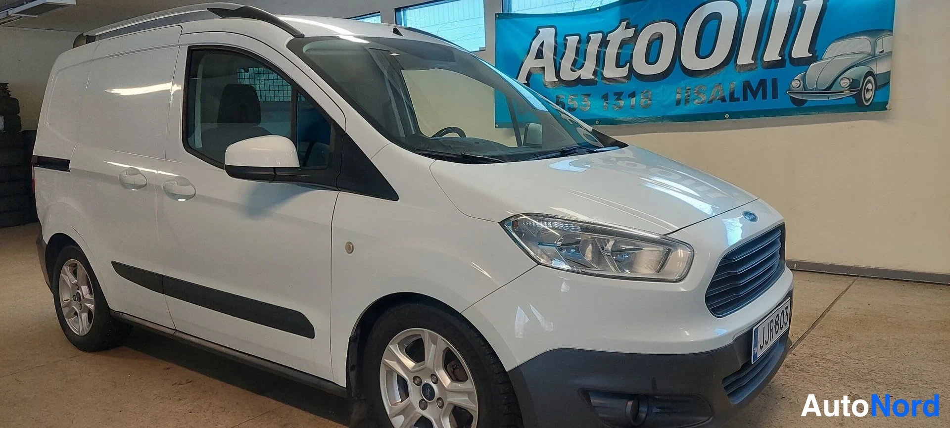 Etuviisto, nokka oikealle: Valkoinen pakettiauto fronttiorieltta, etusäiliö ja kylki näkyvät hallissa.