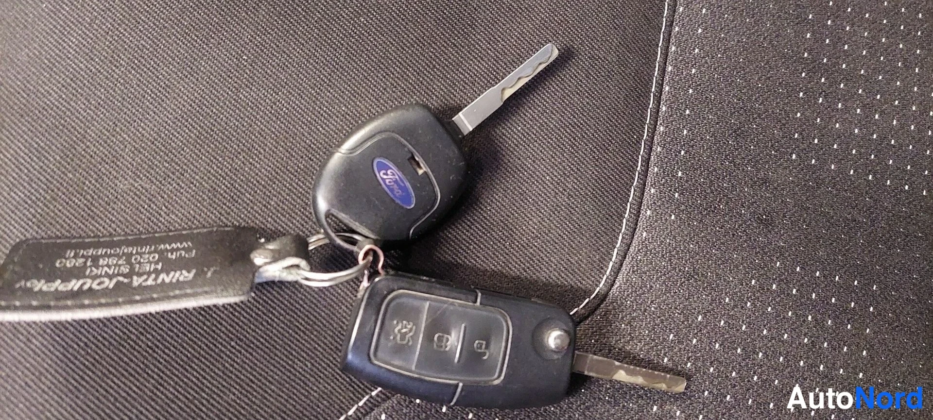 Yksityiskohta: Mustat Ford-avaimet ja kauko-ohjain sinisellä Ford-tunnuksella, tumman harmaalla verhoilulla ja Rinta-Jouppi-avainnauhalla.