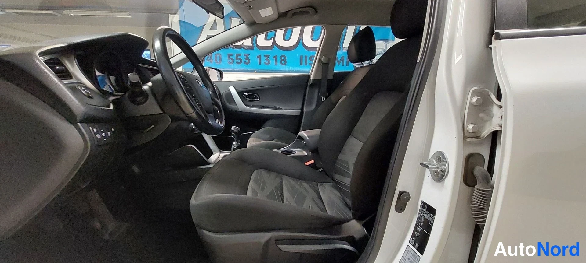 Muu: Kia Ceed Farmari etupenkki ja ohjaamo vasemmalta: musta ohjauspyörä, manuaalivaihteisto ja kangasistuimet; auton runko näkyy valkoisena ovenkarmiin ja B-pylvääs