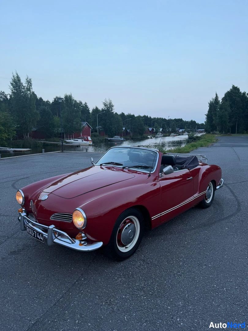 Etuviisto, nokka vasemmalle: Tummanpunainen Volkswagen Karmann-Ghia -cabriolet kolmanneskäänteisessä kuvassa keula vasemmalla, sytytetyt ajovalot ja auki oleva avonainen yläosa.