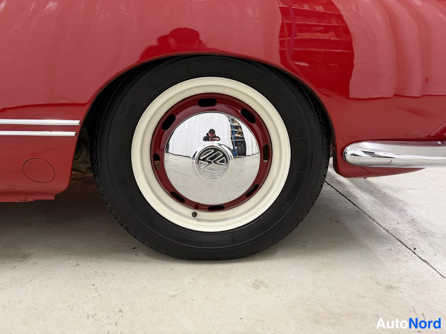 Muu: Punaisen auton pyörä lähikuva; kromi pölykapseli ja valkoreunainen rengas valkoisella korolla, VW-emblem vanteen keskellä. Valkoisessa lattian.