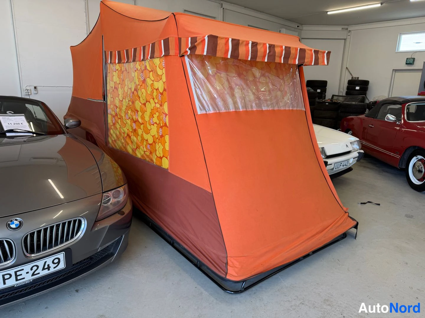 Sivulta: Oranssi telttarakennelma autotallissa; vierellä harmaa BMW-urheiluauto (PE-249) ja taustalla punainen vanha avoauto.