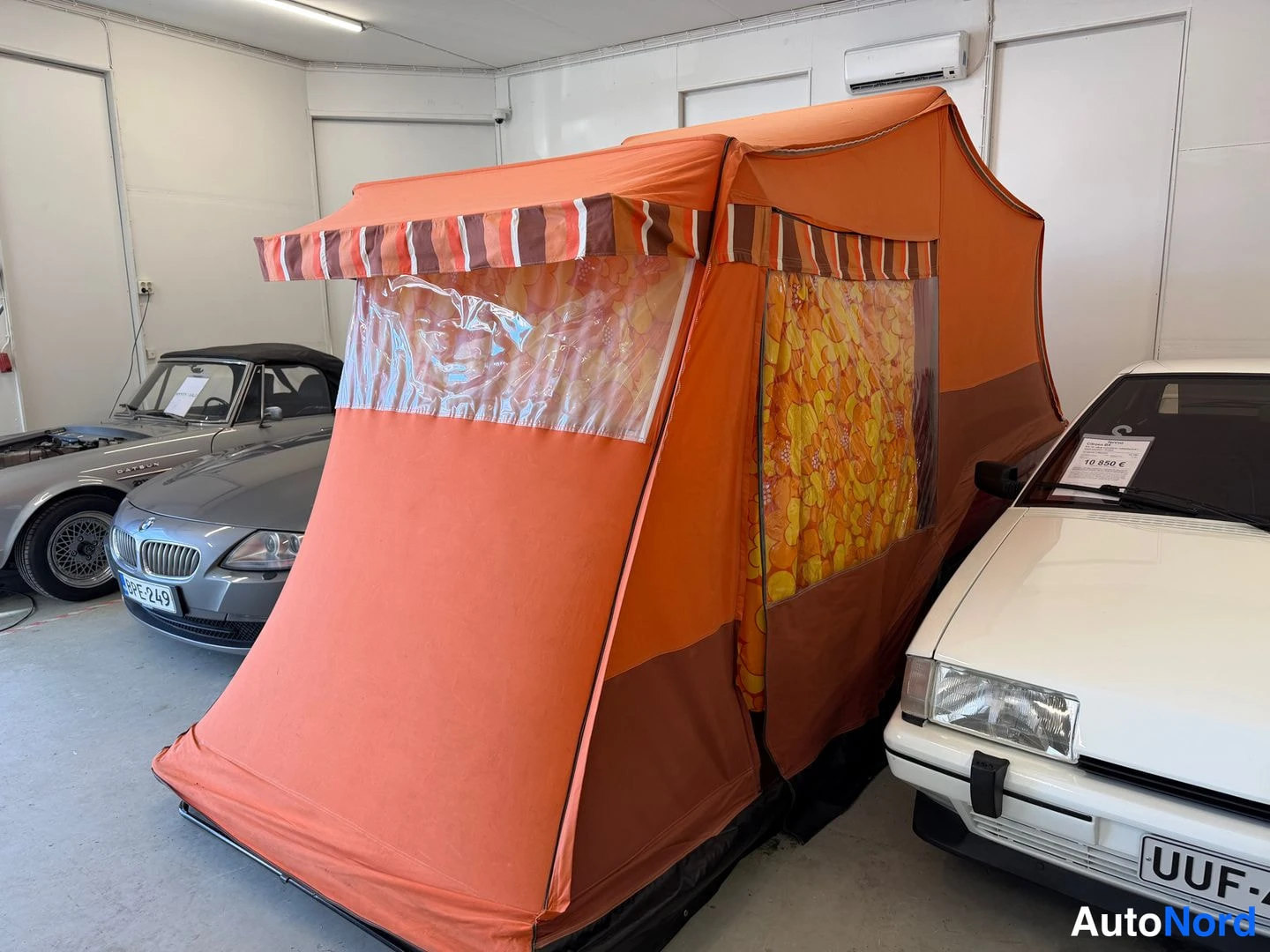 Muu: Oranssi katto- tai telttarakennelma auton takana autotallissa, vierellä valkoinen auto ja harmaita urheiluautoja