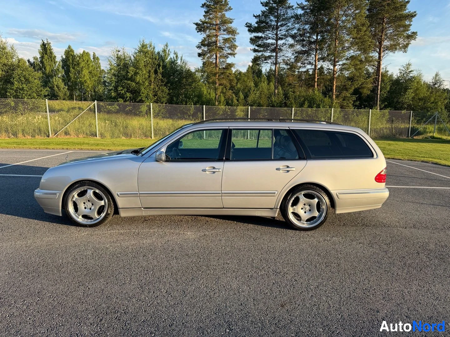 Sivulta: Hopeanvärinen Mercedes-Benz farmariauto sivulta kuvattuna parkkipaikalla puiden ja aitaustaustaa vasten.