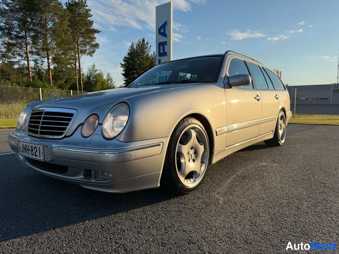Etuviisto, nokka vasemmalle: Farmarimerkki hopeanvärinen Mercedes-Benz farmari etuvasemmasta kulmasta, rekisteritunnus.