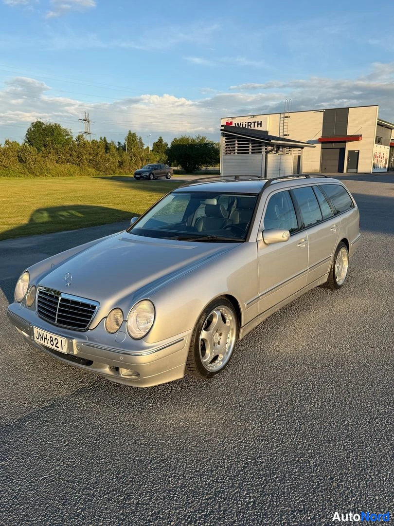 Etuviisto, nokka vasemmalle: Farmarimerkki hopeanvärinen Mercedes-Benz farmari etuvasemmasta kulmasta, rekisteritunnus.
