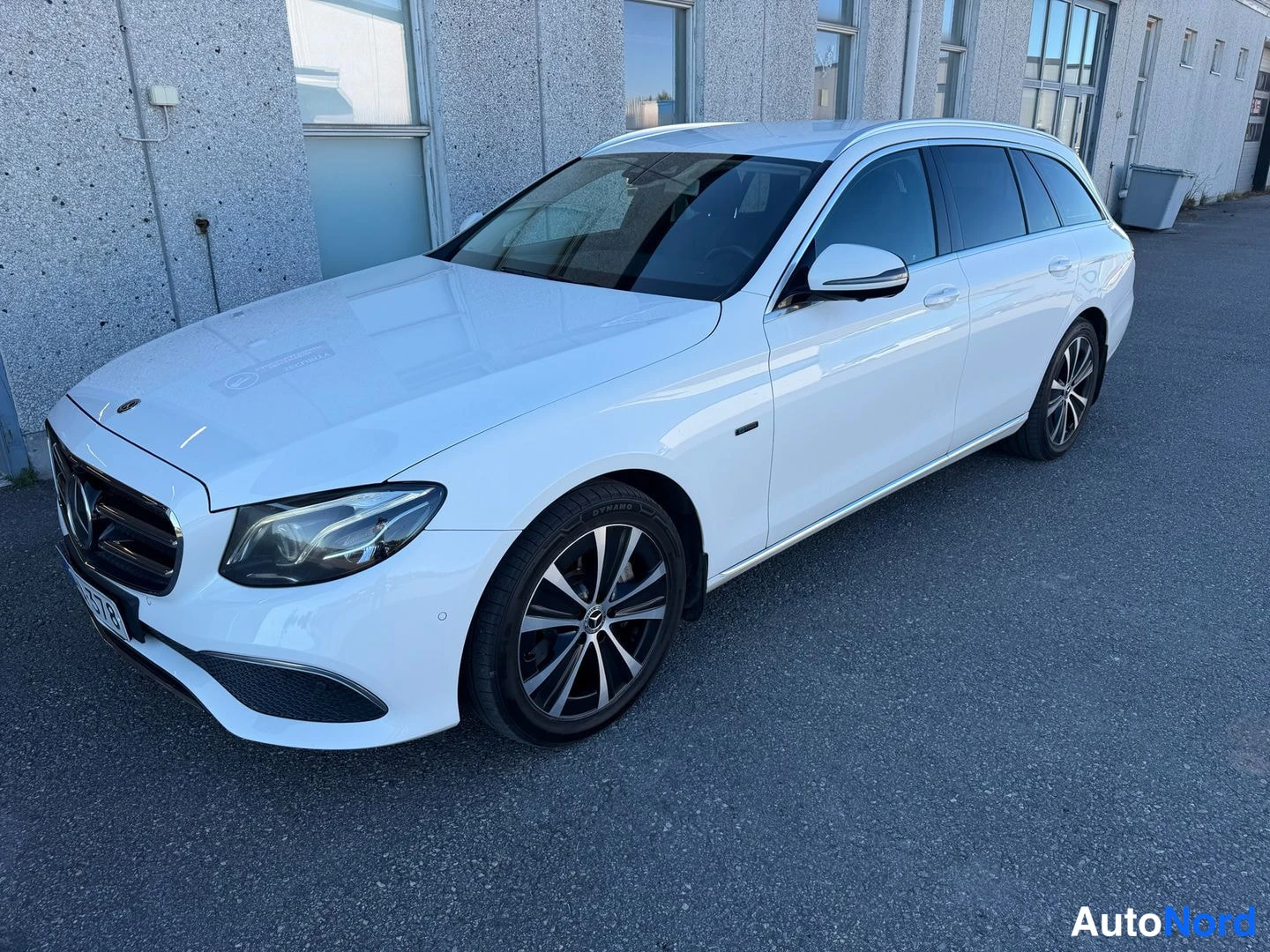 Etuviisto, nokka vasemmalle: Valkoinen Mercedes-Benz E-sarjan farmari kolmasosan etukuva keula vasemmalla, betoniseinä taustalla.
