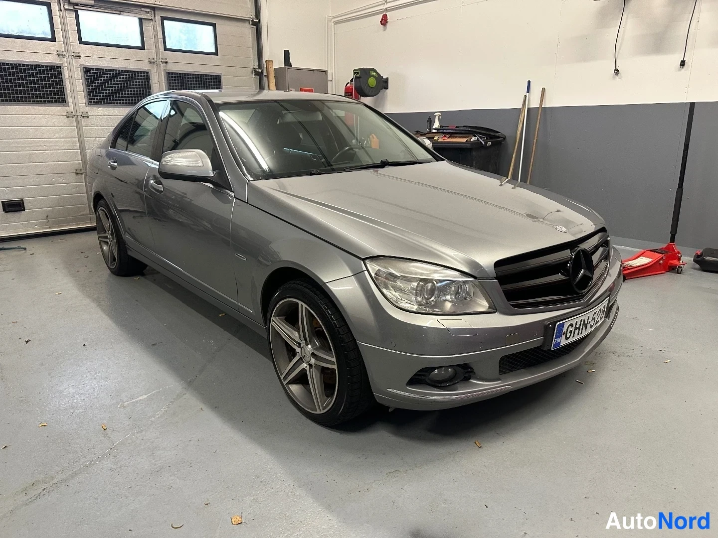 Etuviisto, nokka oikealle: Hopeanharmaa Mercedes-Benz-sedan kulmakuva etupää ja vasen sivu näkyvät auton ollessa tallissa.