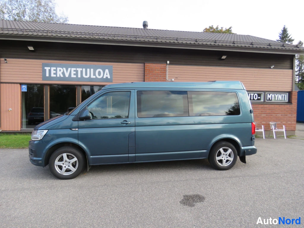 Etuviisto, nokka vasemmalle: Sinertävänharmaan Volkswagen Transporter -pakettiauton sivuprofiili liiketalon edessä, kylki ja keulan etuosa näkyvät.