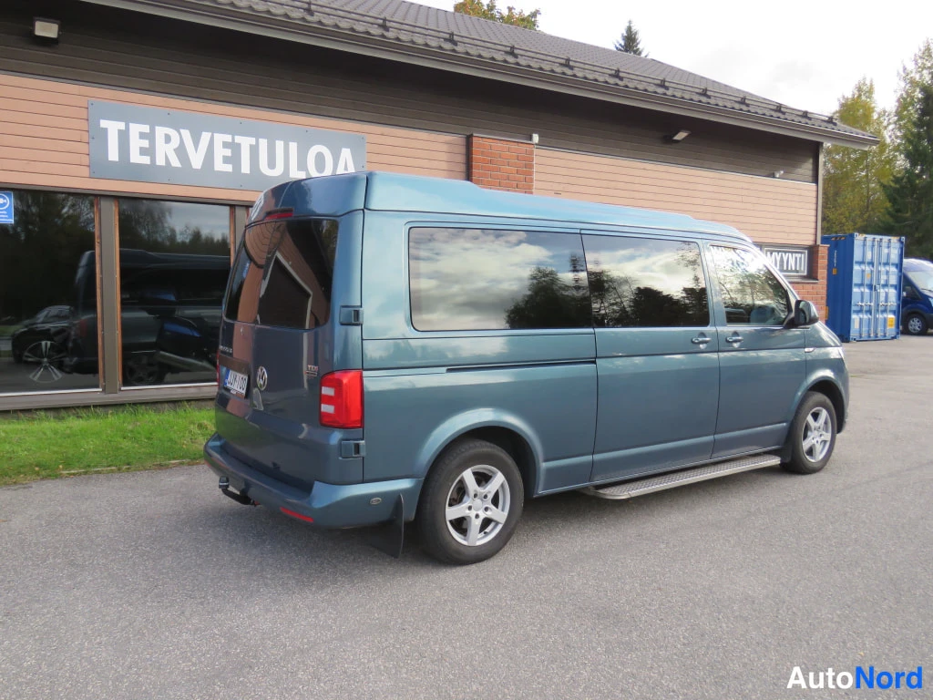 Takaviisto: Sinertävänharmaan Volkswagen Transporter -pakettiauton takaosa ja kylki liiketalon edessä, takavalo ja rekisterikilpi näkyvät.