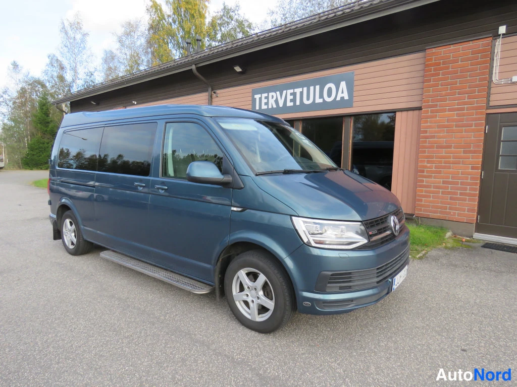 Etuviisto, nokka oikealle: Sinertävänharmaa Volkswagen Transporter -pakettiauto nurkkaetukulmassa talon edustalla, kylkijalkalistat ja entisöity korkea katto näkyvät.