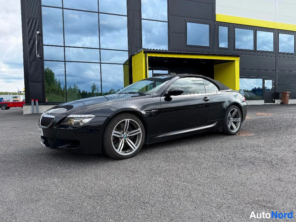 Etuviisto, nokka vasemmalle: Musta avoauto BMW sivukulmassa rakennuksen edessä