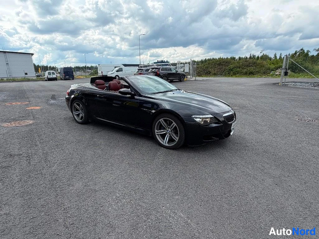 Etuviisto, nokka oikealle: Musta avomallinen urheiluauto kolmasosan etunäkymässä, keula oikealla, aukiolla pilvisen taivaan alla.