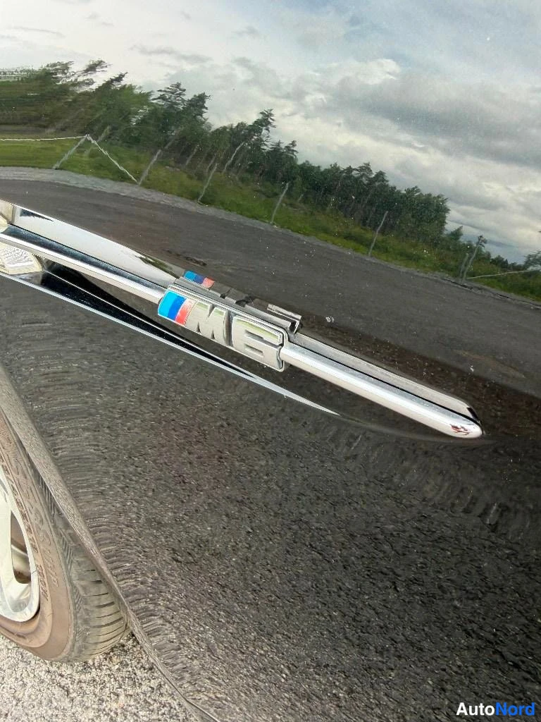 Yksityiskohta: Kromattu M6-tunnus pidikkeessä auton takakannella, kulmikuva.
