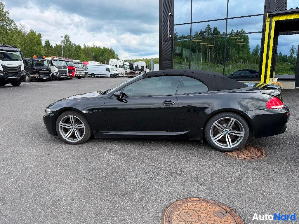 Sivulta: Black BMW M6 convertible photographed from a side profile with the hardtop roof closed, parked on an asphalt lot. Visible features include multi-spoke alloy whe
