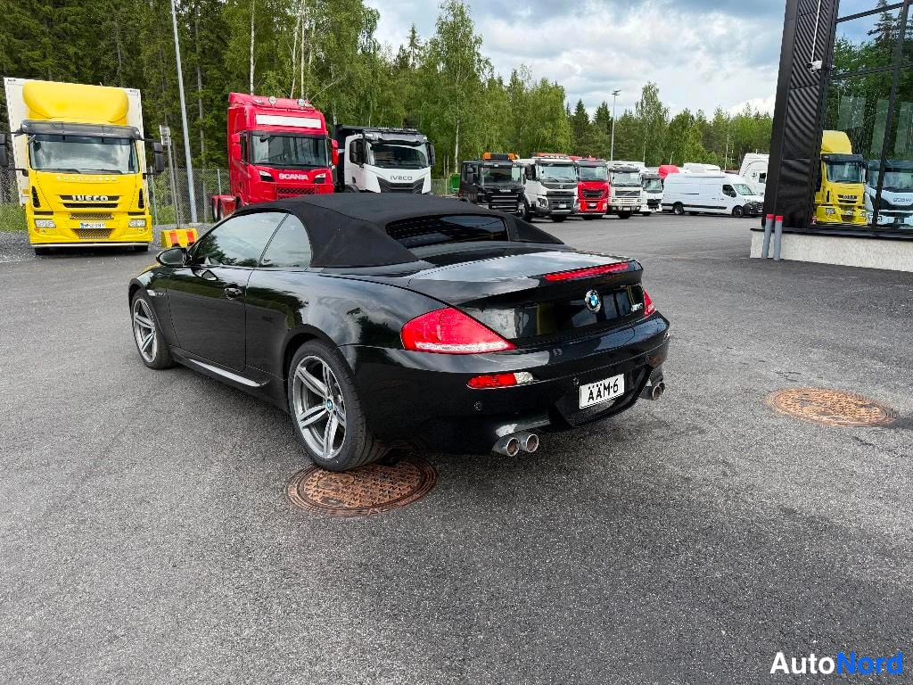 Takaviisto: Rear three-quarter view of the black convertible with the trunk lid showing 'M6' badging and quad exhaust tips. License plate reads ''. Background shows commerc