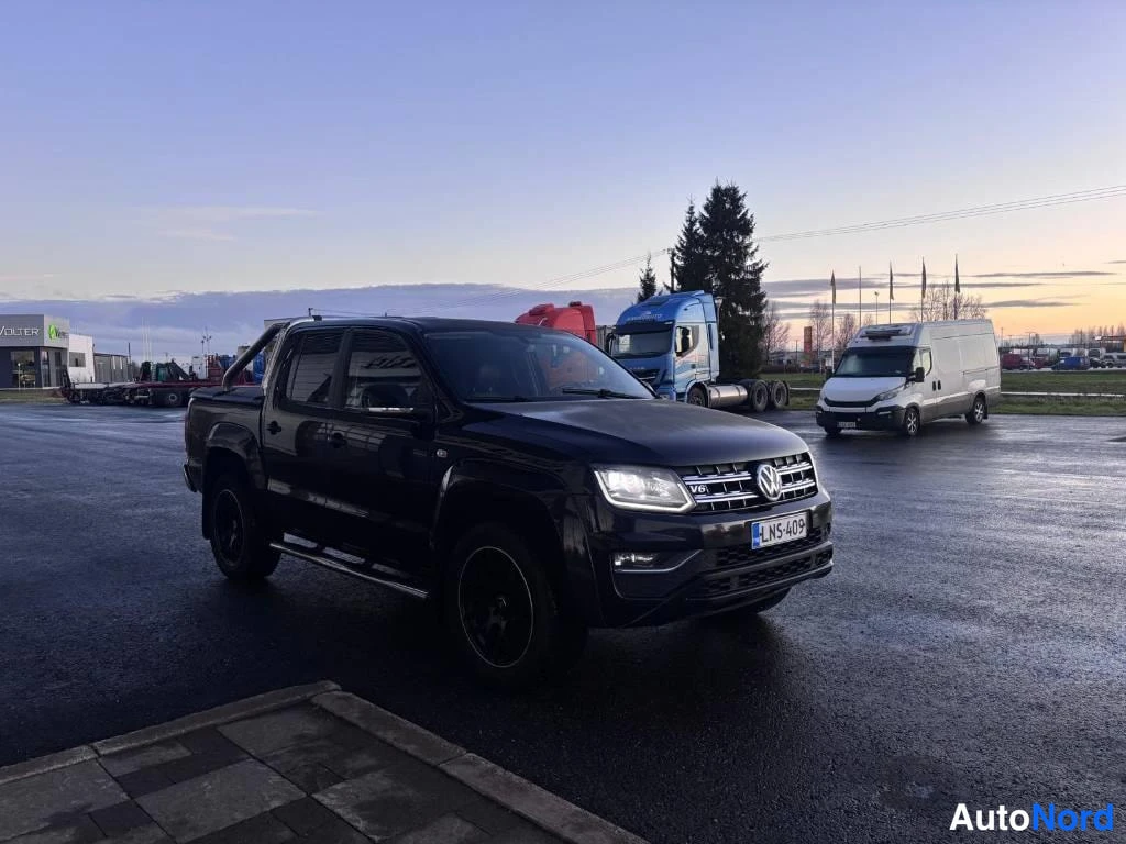 Etuviisto, nokka oikealle: Tummansininen noutoauto pysäköitynä parkkipaikalla illan valossa, keula oikealle.