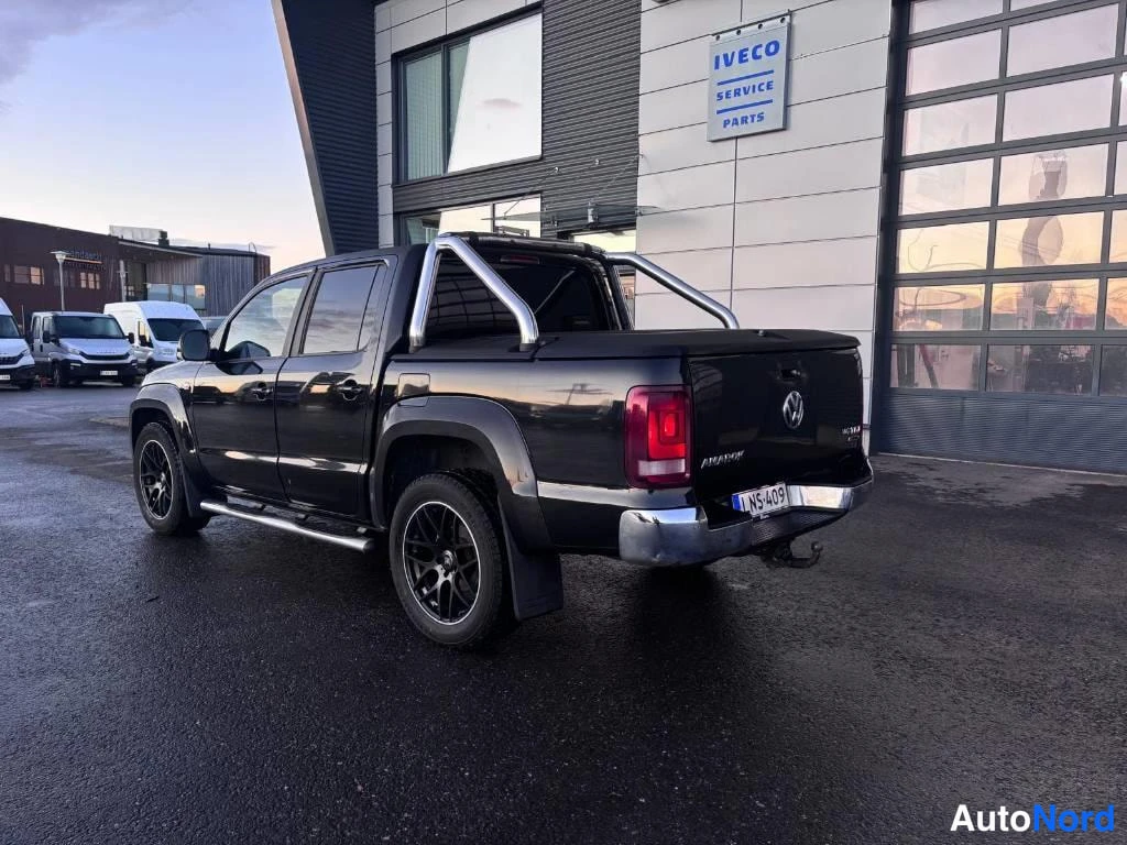 Takaviisto: Musta Volkswagen Amarok -tyynyauto takaa kolmanteen kulmaan nähtynä. Taustalla rakennus, jossa IVECO SERVICE PARTS -mainos. Autossa hopeat metalliset putket tak