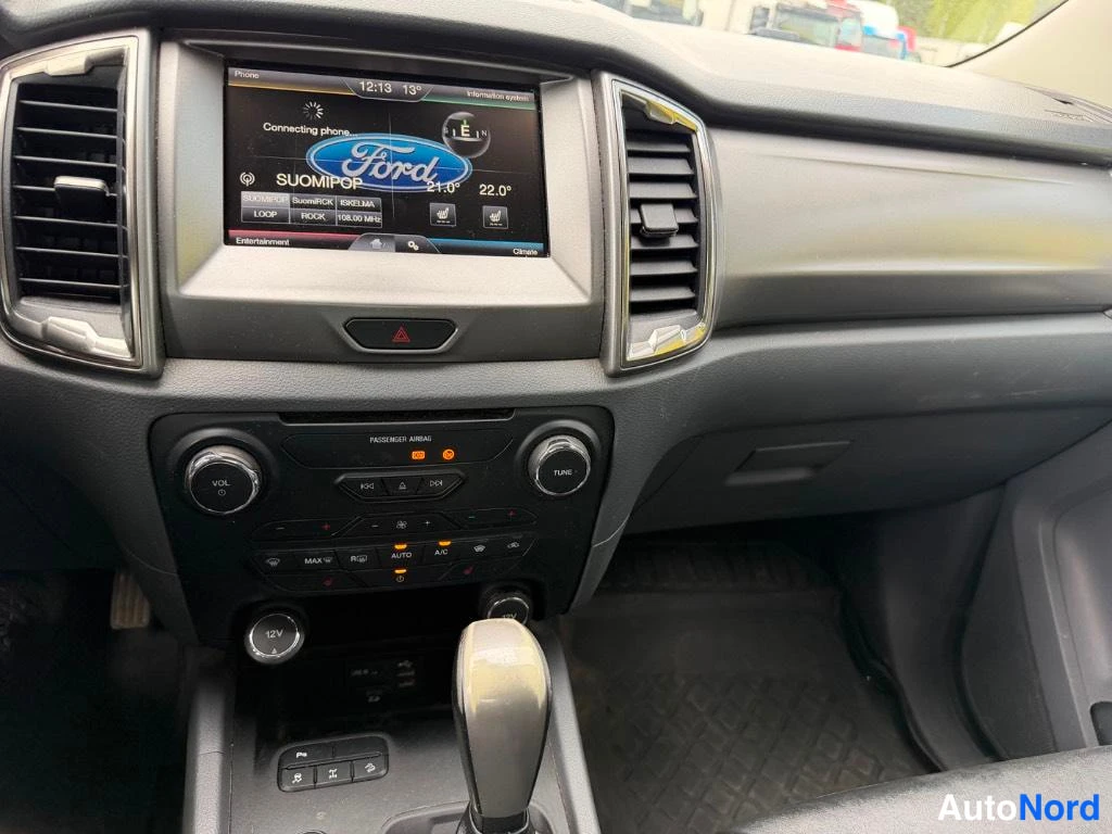 Yksityiskohta: Kohtisuora kuva Fordin kojelaudan keskustasta: kosketusnäyttö näyttää Ford-logon ja radiokanavan SUOMIPOP, alla ilmastointipaneeli A/C- ja AUTO-napeilla, VOL- j