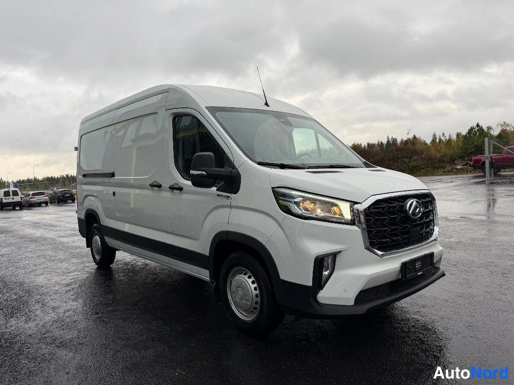 Etuviisto, nokka oikealle: Valkoinen Maxus Deliver 9 -pakettiauto etuviistosta oikealta, keula oikealla, sateisella parkkipaikalla.