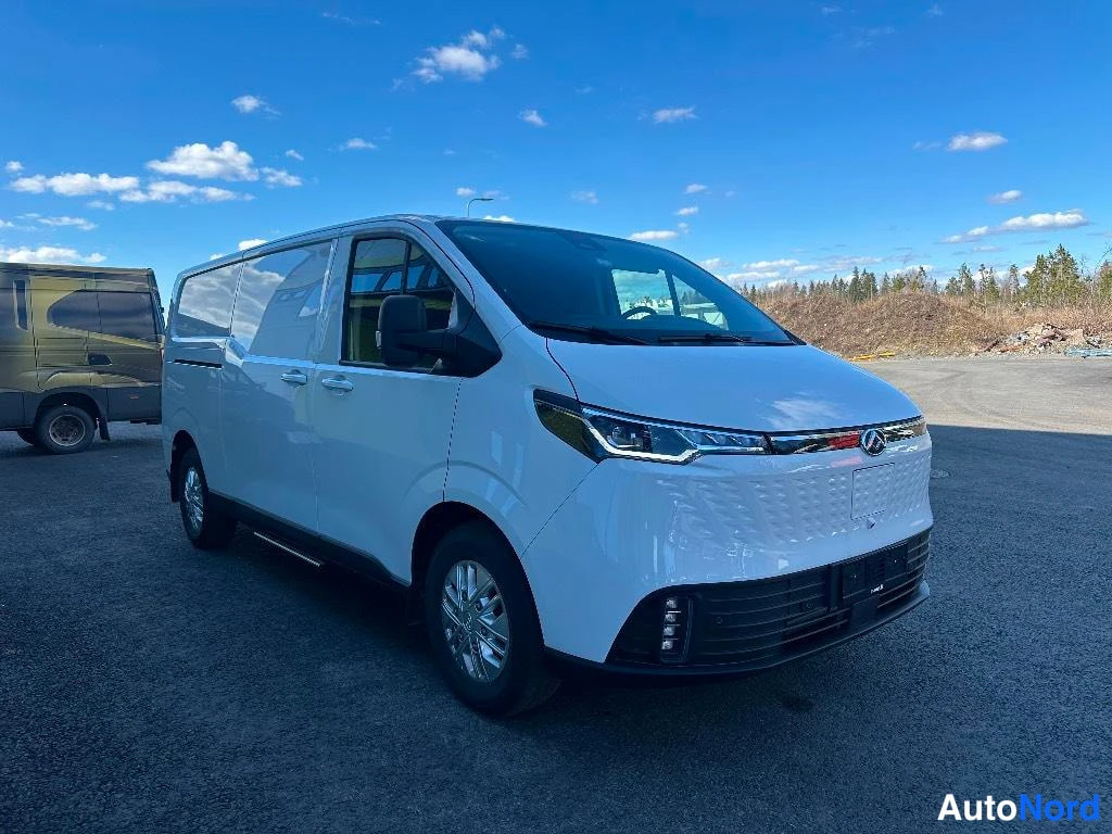 Etuviisto, nokka oikealle: Valkoinen Maxus-pakettiauto etuviistosta oikealta, keulan ja vasemman kyljen näkyessä avoimella piha-alueella.