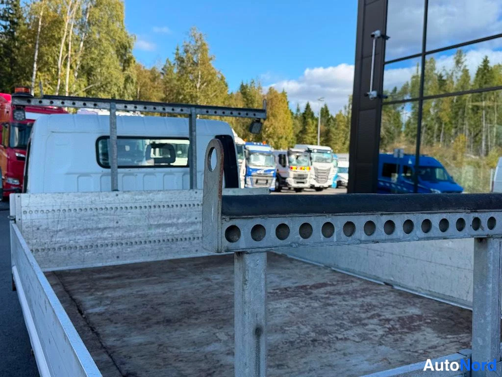 Yksityiskohta: Auton lava-alusta harmaalla kotelolla ja mustalla suojakaiteella, taustalla pysäkkipaikalla muita autokuljetuksia.