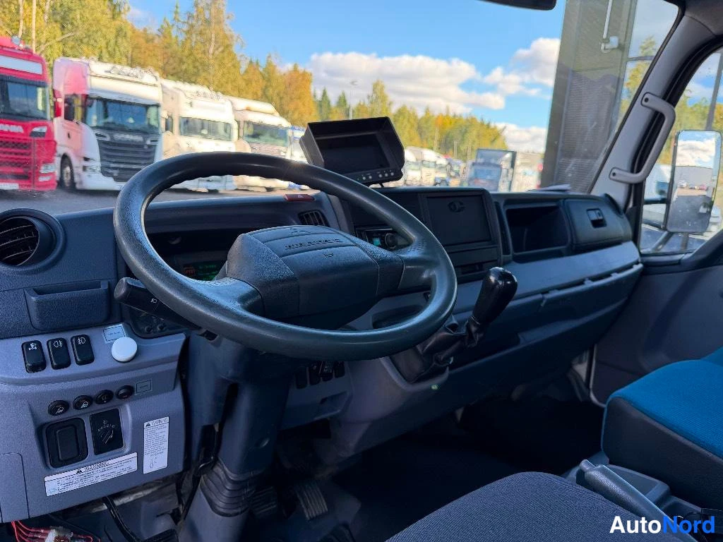 Muu: Kuorma-auton kuljettajan paikka: ratit ohjauspyörä, kojelauta näytöllä ja vaihdevipu, taustalla parkissa kuorma-autoja.