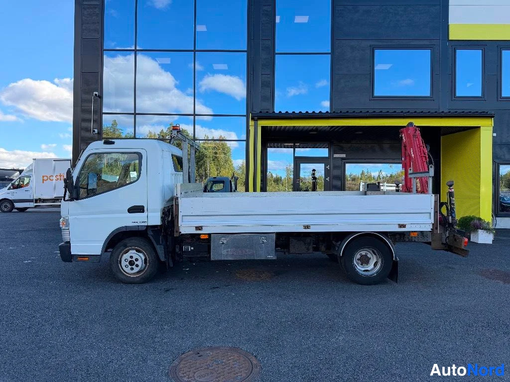 Sivulta: Valkoisen Fuso-avolavan sivuprofiili vasemmalta, alumiinilava, punainen nosturi lavan perässä, taustalla lasipintainen rakennus.